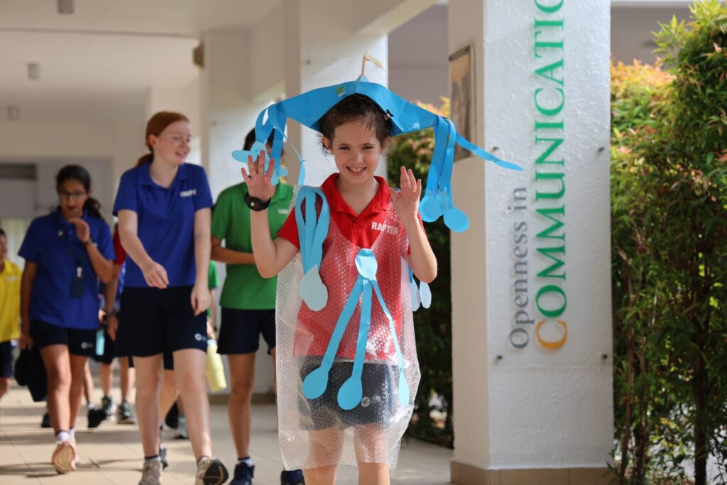 Student wearing a creative costume made from recycled materials walks during a school walkathon at AISM, smiling and raising her hands.