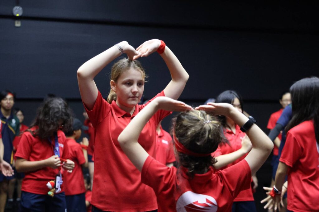 Students in red sports uniforms practice coordinated movements during a team-building or mindfulness activity indoors at AISM.
